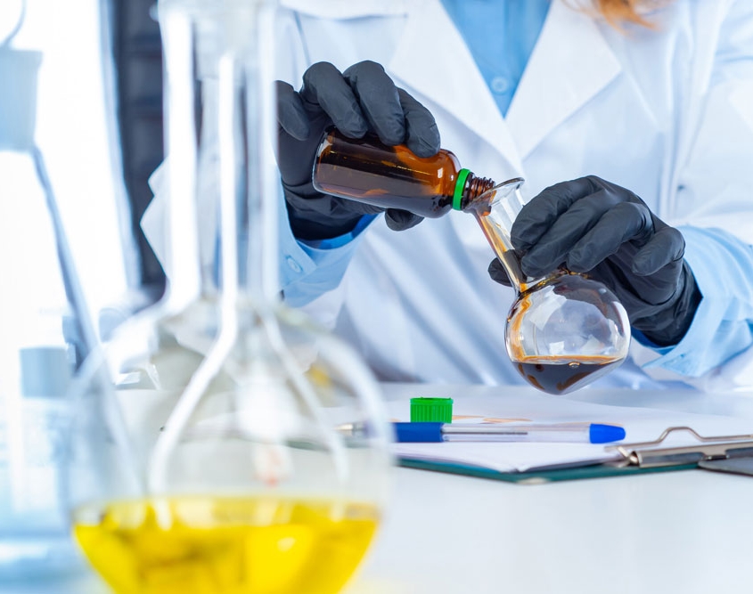 Woman in lab coat pouring brown chemical into a glass beaker.